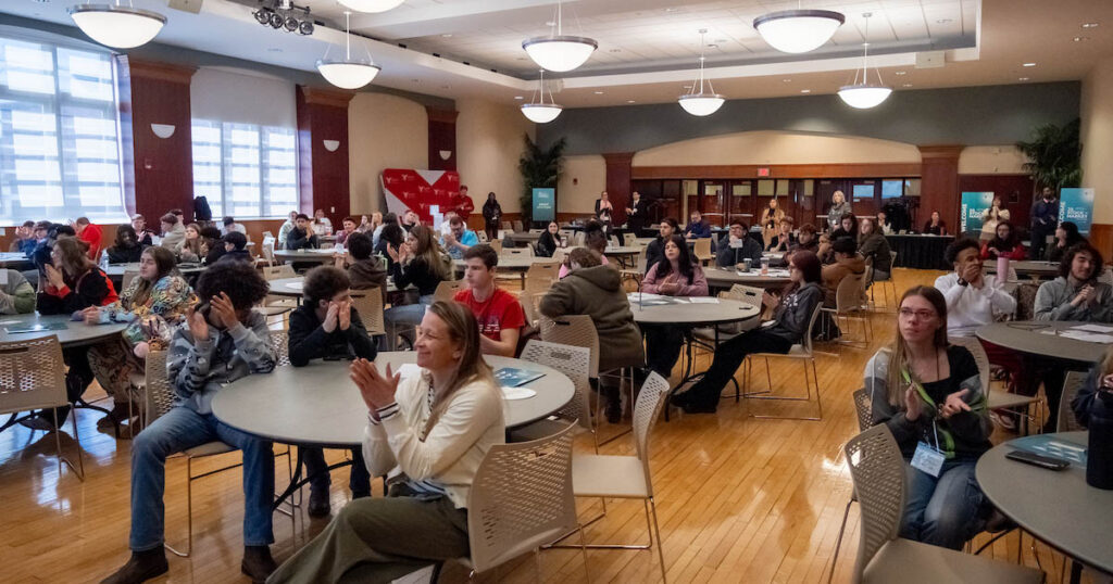 People seated at various tables in a large, well-lit room smile and clap at an event.