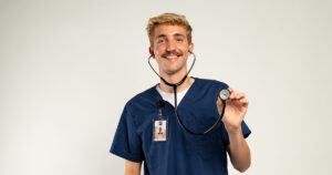 A young man with short blonde hair wearing a blue scrub top and a stethoscope, posing and smiling in front of a white backdrop.