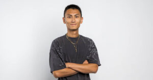 Portrait of Reo Horie, wearing a black t-shirt and gold necklace while smiling and crossing his arms.