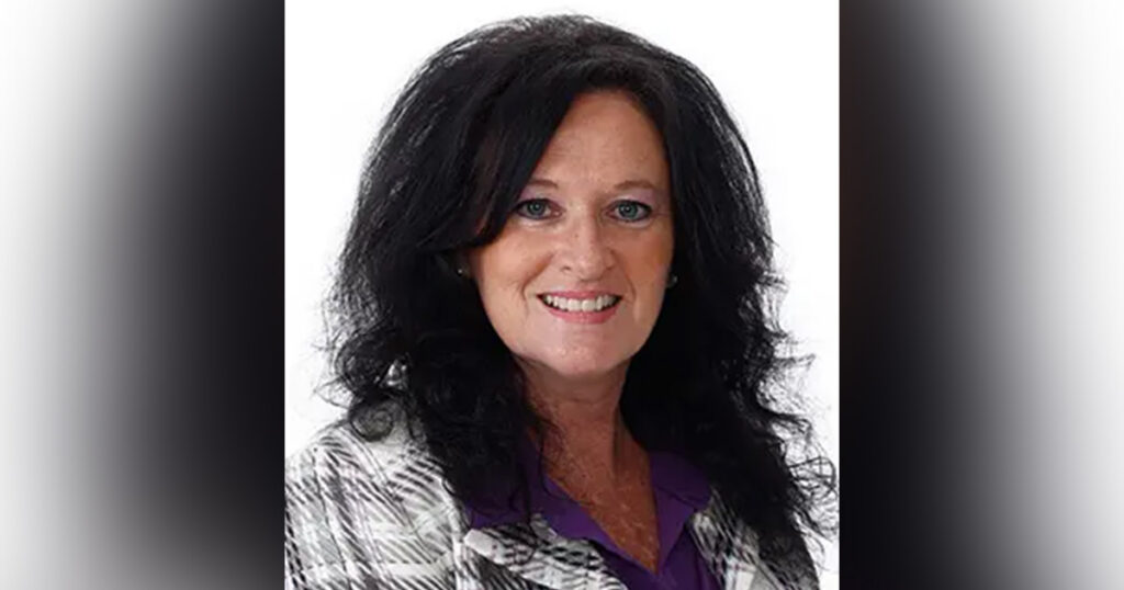 Dixie Denton, a woman with medium-length black hair smiles and poses while wearing a white and gray plaid jacket and a purple shirt.