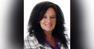 Dixie Denton, a woman with medium-length black hair smiles and poses while wearing a white and gray plaid jacket and a purple shirt.