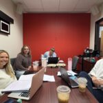 Four women at a conference table with laptops, notebooks, and drinks. Three smile at the camera; one focuses on a laptop. Red wall background.