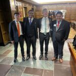 Four men in suits wearing orange, blue, green, and red ties with Ball State cardinal lapel pins stand smiling in a wood-paneled lobby.
