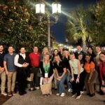 A group of 20 smiling adults, men and women, pose outdoors at night under a two-lamp streetlamp. Trees and buildings are visible in the background.