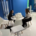 Two women sit at a white table with multiple rolls of toilet paper, coffee cups, and a phone, conversing. Blue walls form the background.