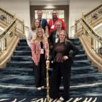 Six individuals pose on a grand staircase with patterned blue carpet and ornate gold railings. All wear event lanyards and badges.