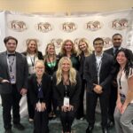 Ten participants in business attire, wearing lanyards, smile in front of an ICSC Sales Competition banner.