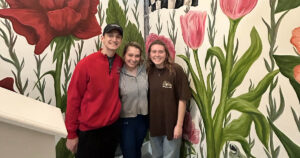 Three students stand and smile in front of an indoor mural that displays various large pink and red flowers with greenery