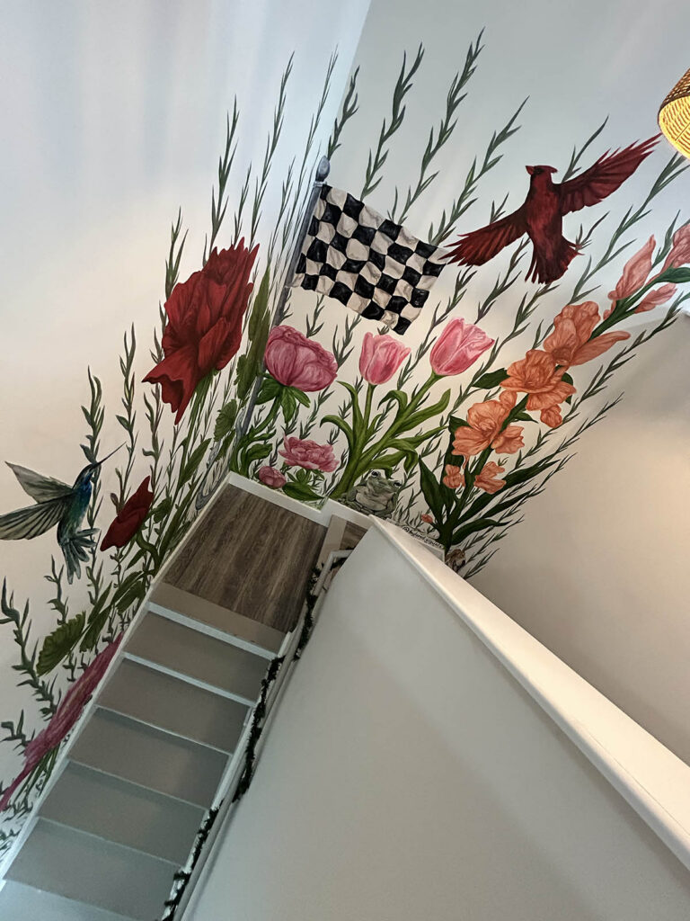 Top-down view of a mural that covers the walls of a staircase landing. The mural is made up of various large pink and red flowers and greenery, with a checkered flag and a red cardinal soaring above.