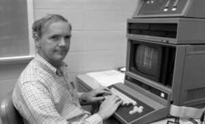 Dr. Paul R. Errington sits at a keyboard beside a computer terminal in a Ball State University lab, reflecting the hands-on electronics work and teaching that defined his 34-year career.