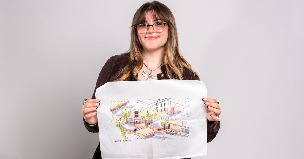 Brooke Wagner, a young woman with long brown hair and bangs wearing glasses ans smiling while holding up a paper with an architectural drawing on it.