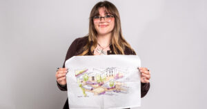 Brooke Wagner, a young woman with long brown hair and bangs wearing glasses ans smiling while holding up a paper with an architectural drawing on it.