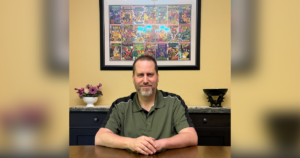 Scott Lantz sitting at a table with a large framed picture of comic books behind him.