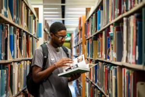 A student looks through a book while surrounded by bookshelves in a library.