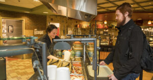 A student holding a tray laughs with a dining worker while waiting in line at a campus dining complex.