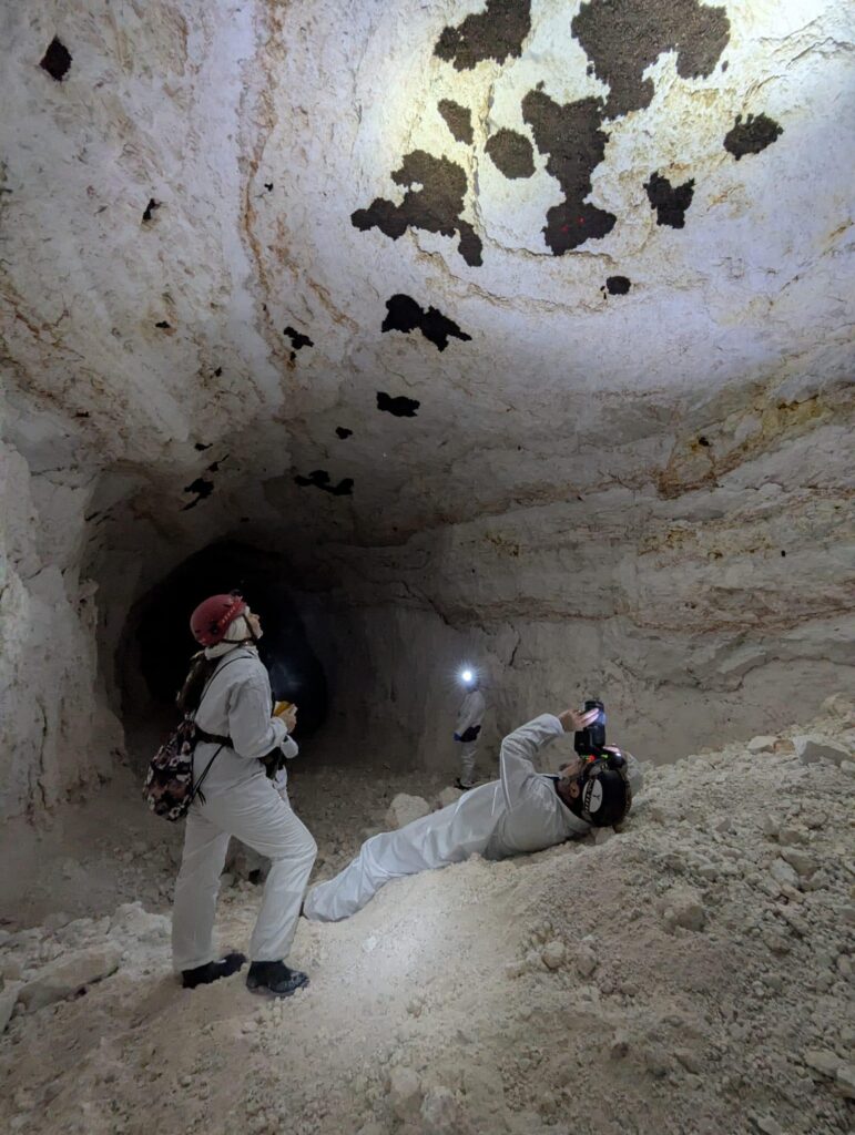 Three people in white coveralls and helmets are in a cave. One person lies on the ground taking a photo of the ceiling, while another stands and looks up.