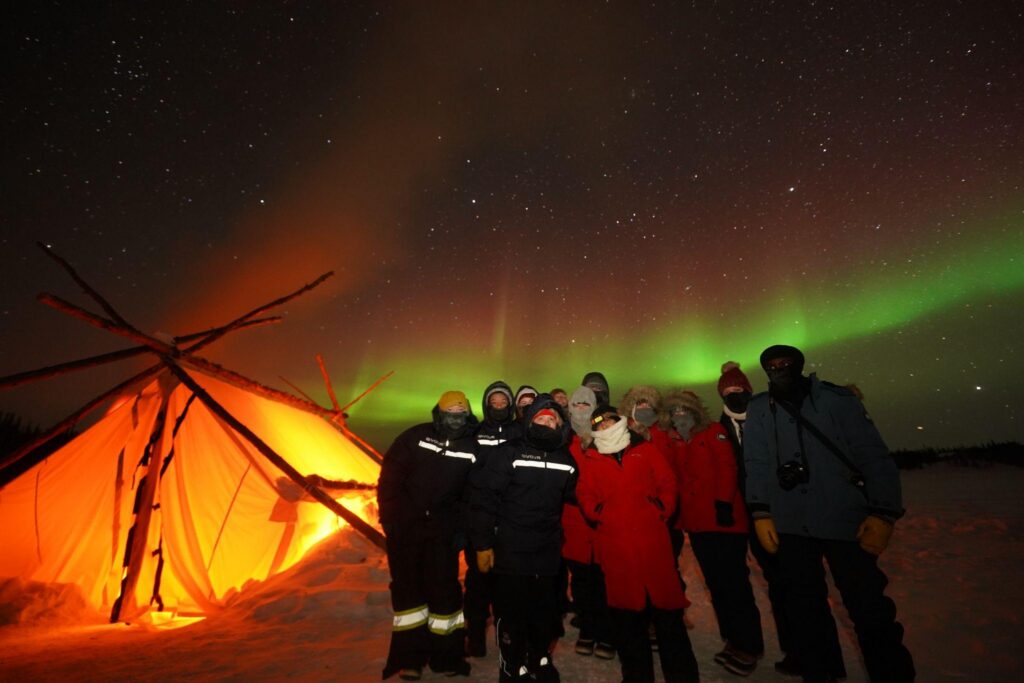 A group of people in winter gear stand in the snow at night under a green and red aurora borealis, next to a glowing orange teepee.