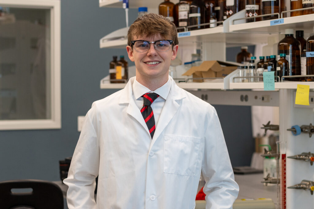 A smiling man with glasses wears a white lab coat over a white shirt and a red and black striped tie, standing in a room with shelves of bottles.