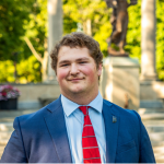 Young man in a blue suit standing in front of a distant Benny statue.