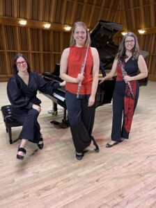 three women on a stage, one seated at a grand piano on the left, one posed standing with a flute in the middle, one posed standing with an oboe