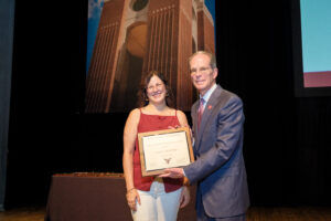 two people on a stage. on the left a brunette caucasian woman receives a plaque from a caucasian man on her right. both are smiling at the camera