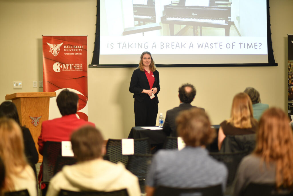 A woman in a black blazer and red shirt stands before an audience, gesturing with her hands. A screen behind her displays a piano and the text, "Is taking a break a waste of time?"