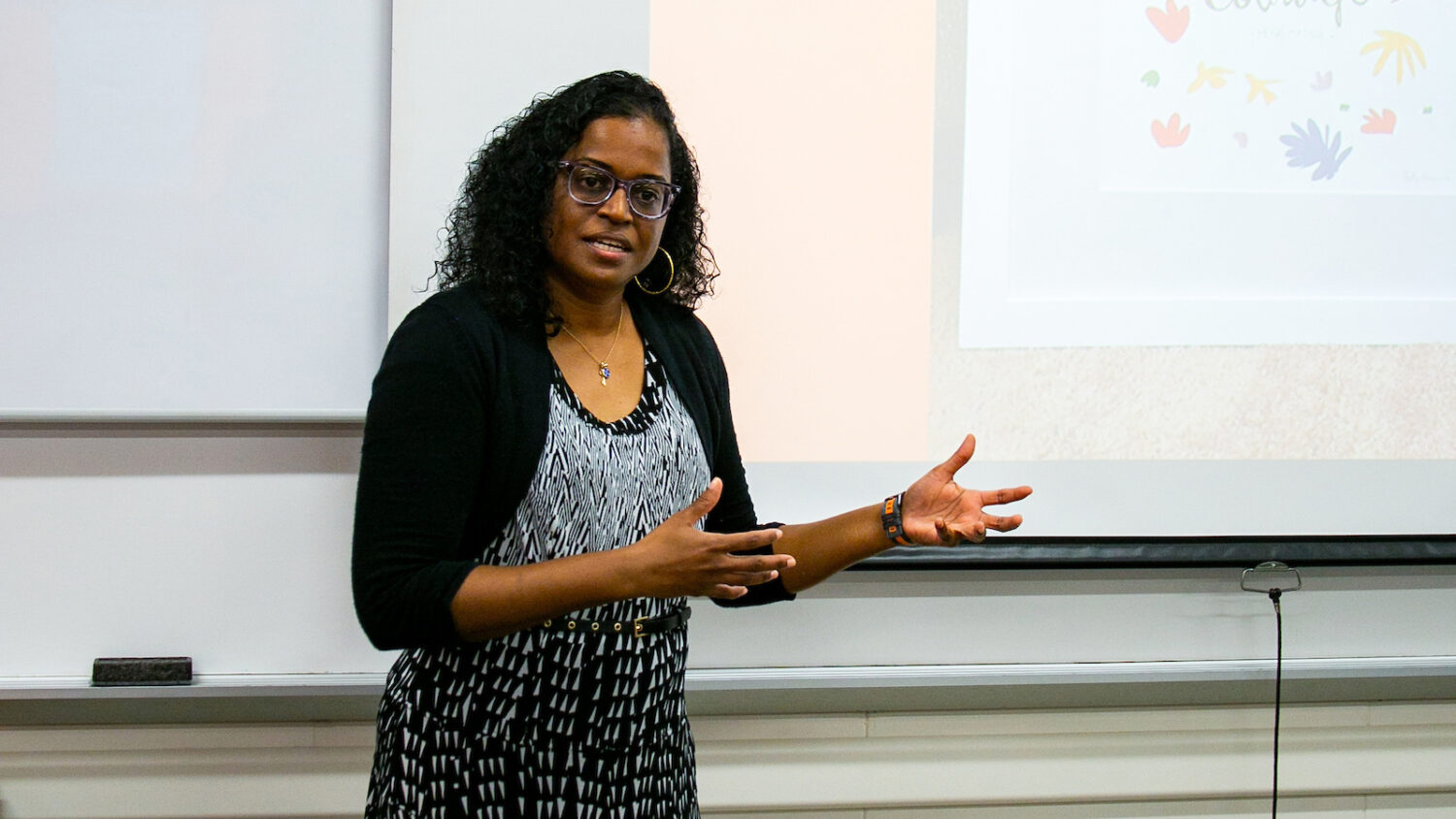 A woman with curly dark hair and glasses wears a black cardigan over a patterned black and white dress, gesturing with her hands while speaking.