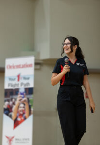 A person presents on stage with a microphone in their hand. A sign with the word orientation is showcased in the background.