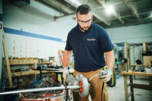A man wearing safety glasses, gloves, and a navy blue polo shirt labeled “electrical training” works with electrical conduit and tools in a workshop filled with wiring, equipment, and wooden shelves.