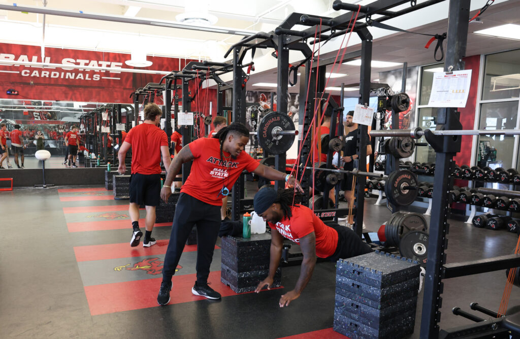 Kam Bryant leans over to coach a Ball State football player doing a push-up in the Cardinals weight room while teammates in red shirts walk behind them.