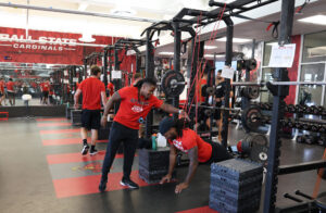 Kam Bryant leans over to coach a Ball State football player doing a push-up in the Cardinals weight room while teammates in red shirts walk behind them.