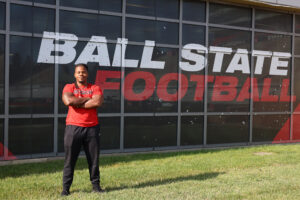Kam Bryant stands with arms crossed on the grass in front of the “Ball State Football” window graphics, wearing a red Attack shirt and black pants. 