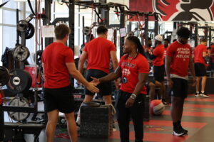 Kam Bryant smiles and reaches out to high-five a Ball State football player in the weight room as teammates train in the background.