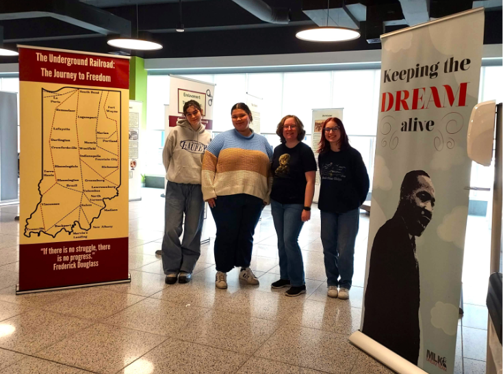 Four students from COMM450 pose with banners at their showcase of their "Journey to Freedom" exhibit.
