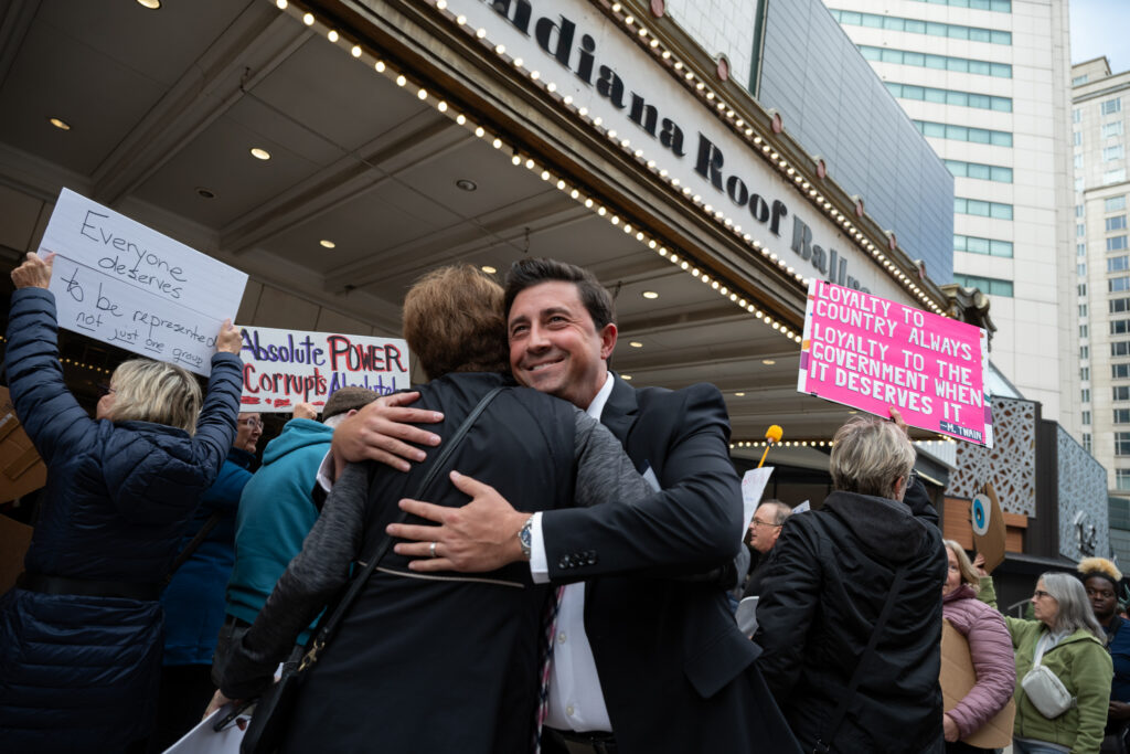A smiling man in a suit embraces a protester outside the Indiana Roof Ballroom in Indianapolis, surrounded by demonstrators holding signs criticizing political power and government accountability. The scene captures a moment of personal connection amid a public protest against mid-cycle redistricting.