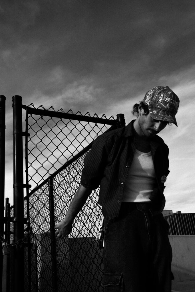 A black-and-white portrait of a young man in a camouflage cap and open over shirt leaning against a chain-link fence, his gaze cast downward beneath a dramatic, cloudy sky.