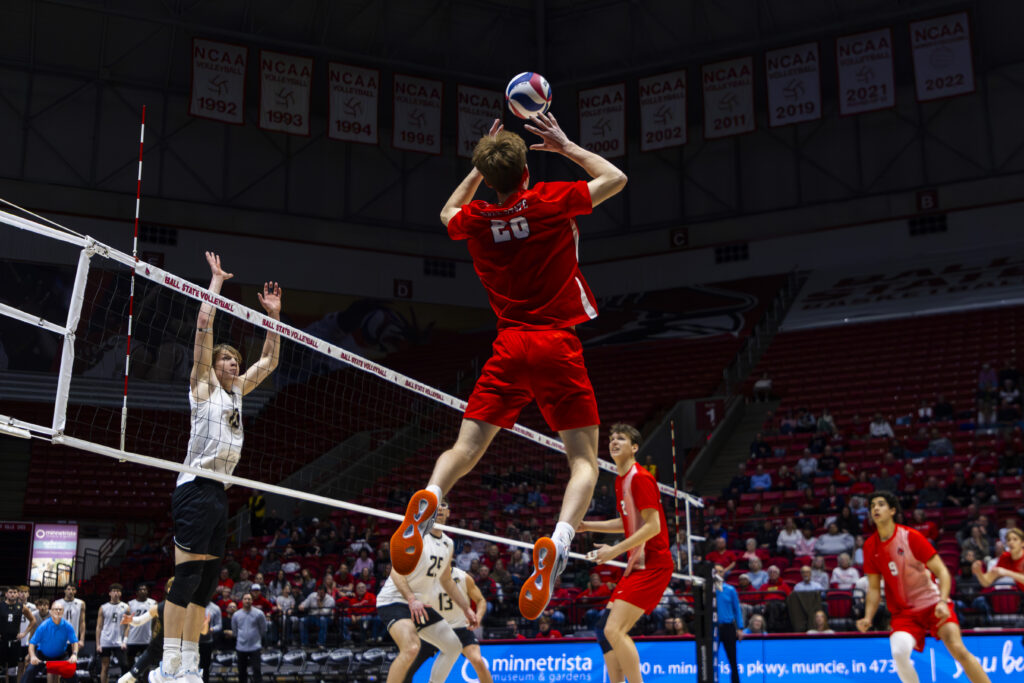 A Ball State volleyball player in red, wearing number 20, leaps above the net to set the ball during a home match, with an opposing player in white reaching up to block. NCAA championship banners from various years line the rafters of the arena above the action.
