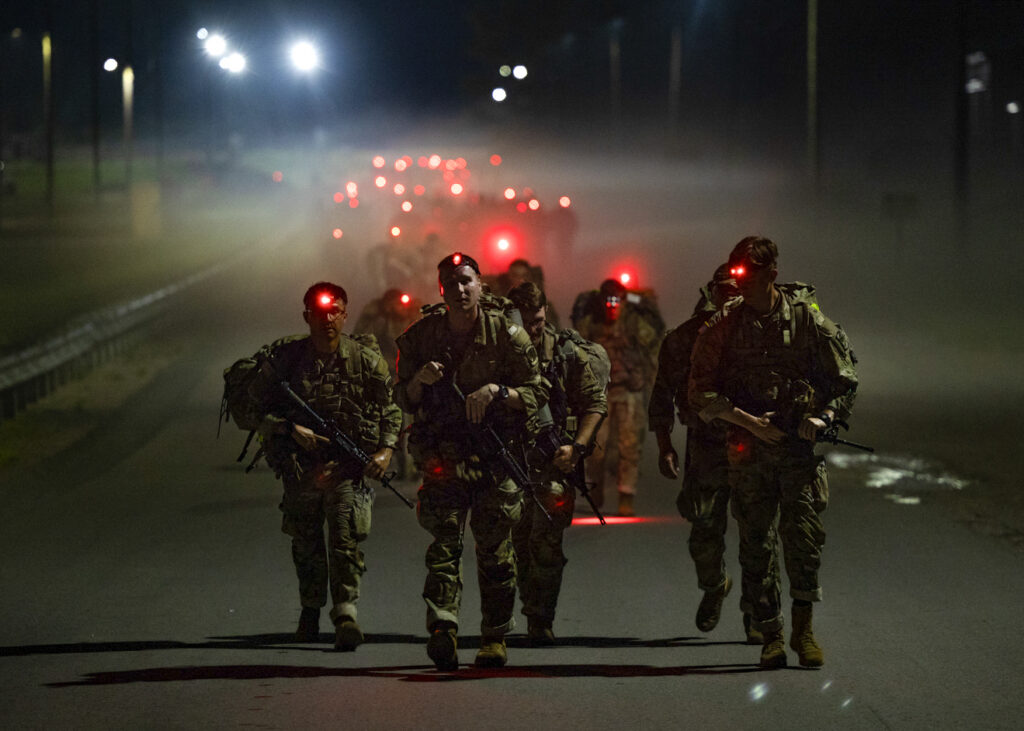 A group of military cadets in full combat gear marches down a dark road at night, their red headlamps glowing through a haze as they carry rifles during a nighttime ruck march. The dramatic scene stretches into the foggy background, where additional cadets and red lights recede into the darkness.