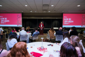 A speaker in a red blazer addresses a banquet room full of attendees seated at round tables during Ball State University's 2026 Louie Awards ceremony. Two large red screens displaying the "Louie Awards 2026" branding flank the podium at the front of the room.