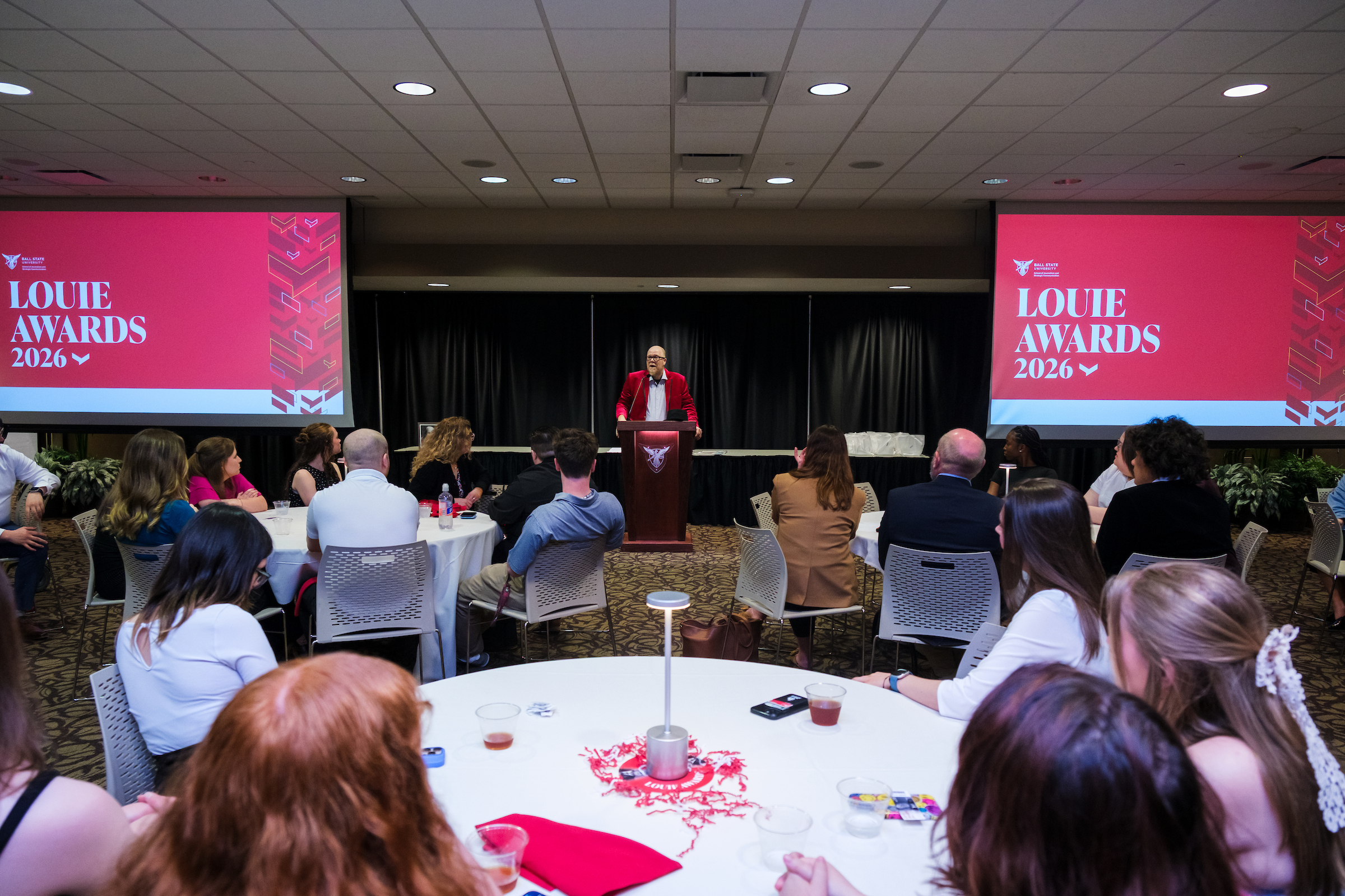 A speaker in a red blazer addresses a banquet room full of attendees seated at round tables during Ball State University's 2026 Louie Awards ceremony. Two large red screens displaying the "Louie Awards 2026" branding flank the podium at the front of the room.