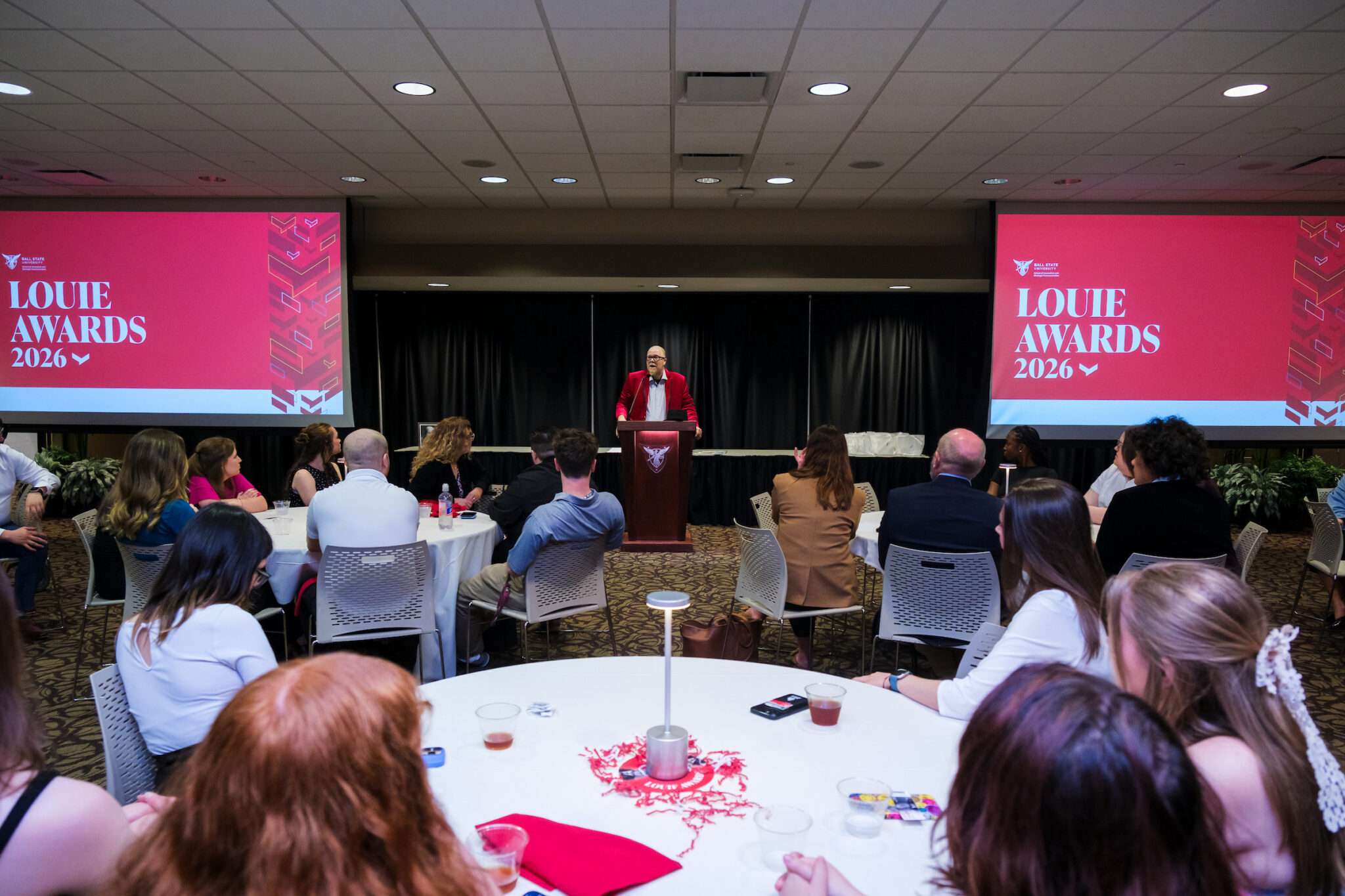 A speaker in a red blazer addresses a seated audience at the Louie Awards 2026 ceremony, with two large pink projection screens displaying the event name flanking the podium.