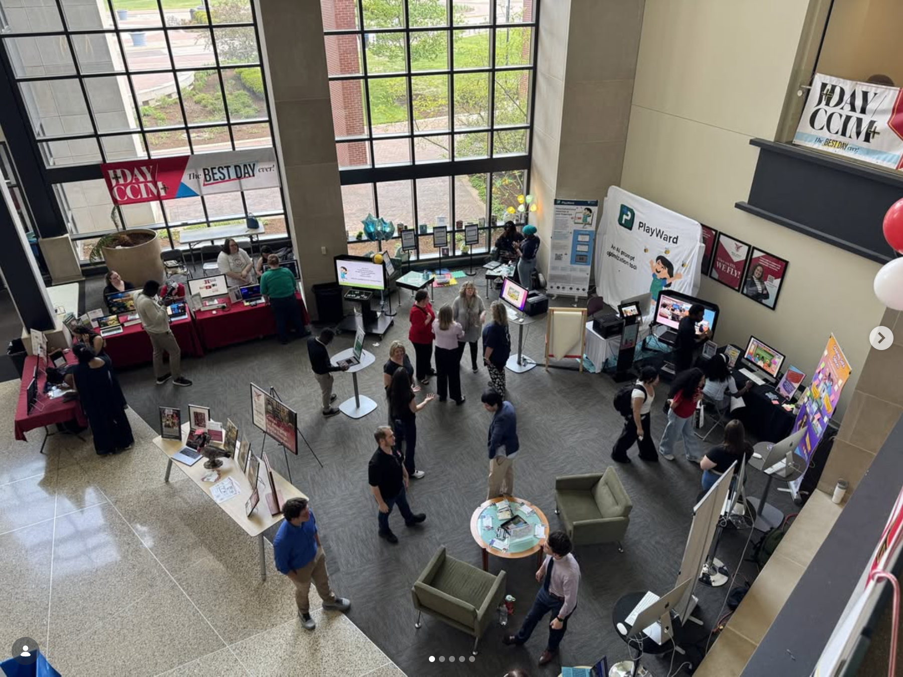 An overhead view of a busy indoor event or expo in a modern building atrium, with attendees browsing multiple vendor or student organization tables and displays set up across the floor.