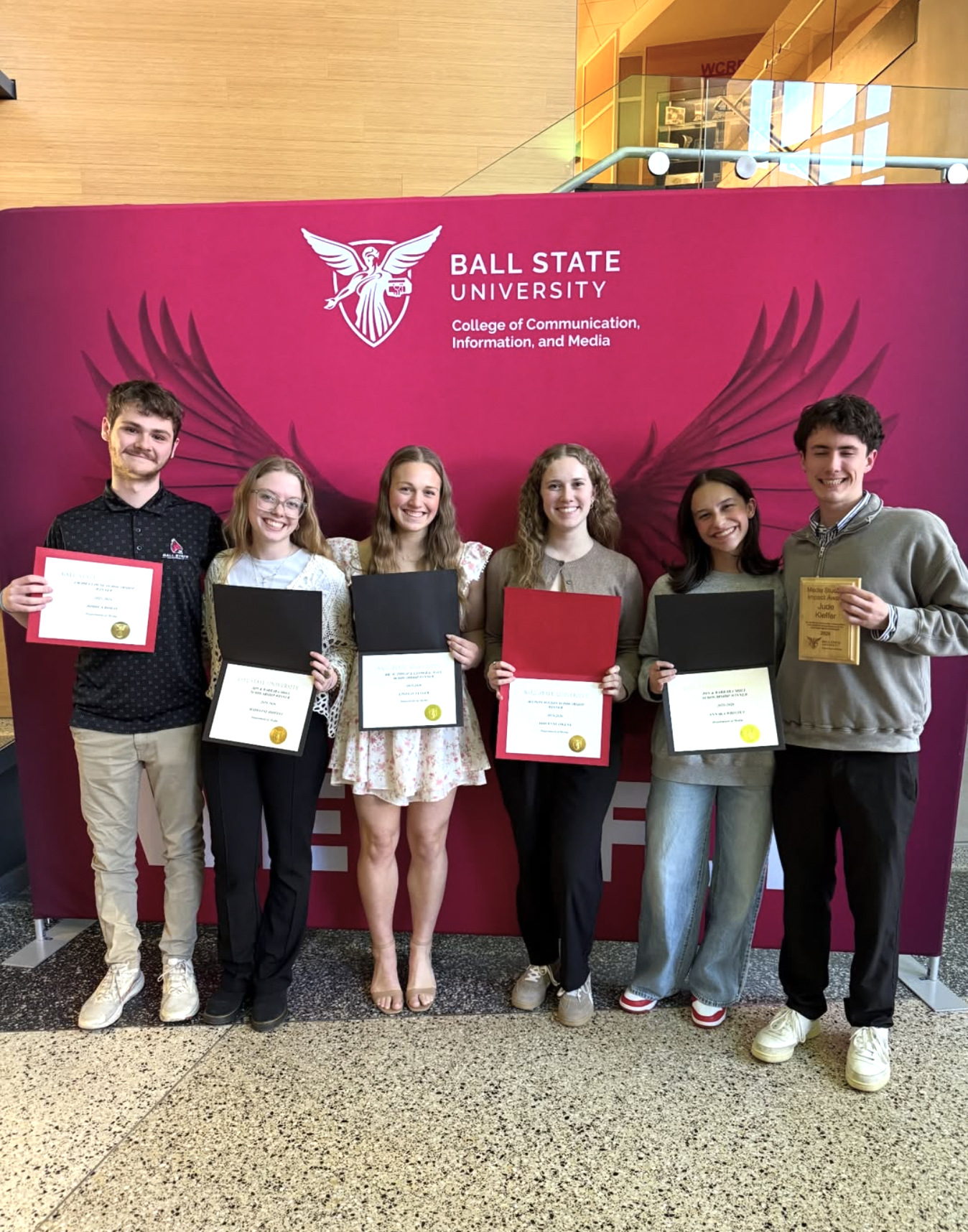 Six smiling students hold awards and certificates in front of a Ball State University College of Communication, Information, and Media backdrop, celebrating their achievements at a recognition event.