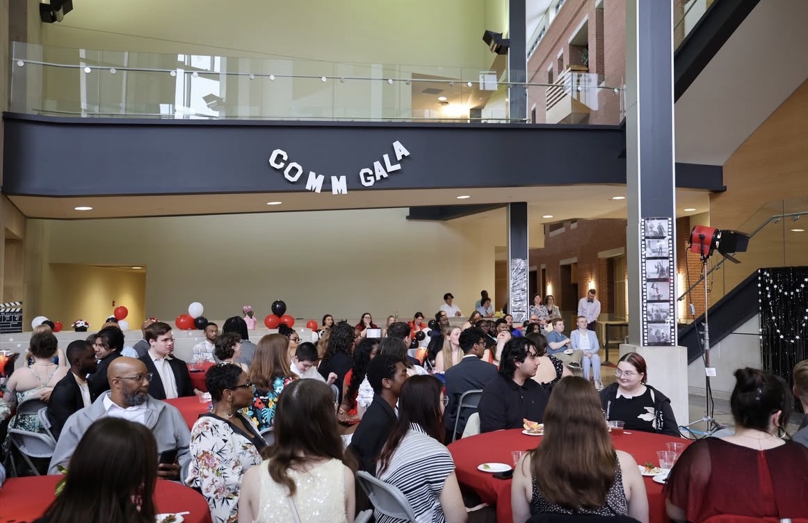A large crowd of students and guests are seated at round tables with red tablecloths during the Comm Gala, held in a modern building atrium decorated with red and black balloons and a film strip display.