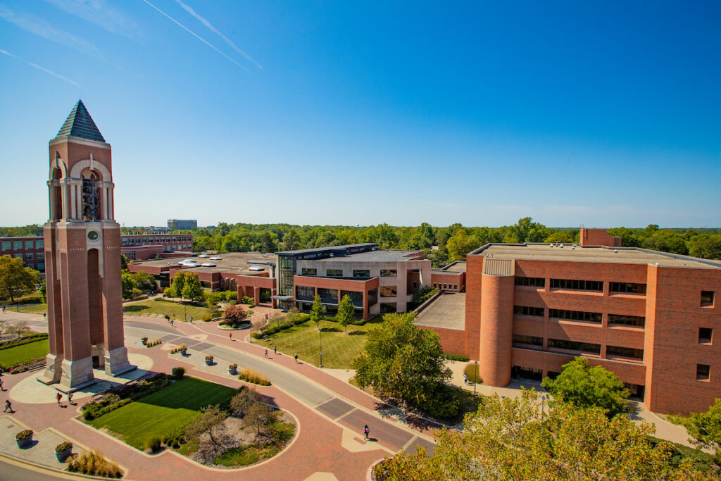 An aerial view of Ball State University's campus on a sunny day, featuring the iconic Shafer Tower bell tower on the left, surrounded by brick walkways, green lawns, and several multi-story academic buildings stretching into a tree-lined horizon.
