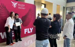 Two people pose for a photo in front of a red backdrop with wings while two others closer in the foreground of the image take their photo.