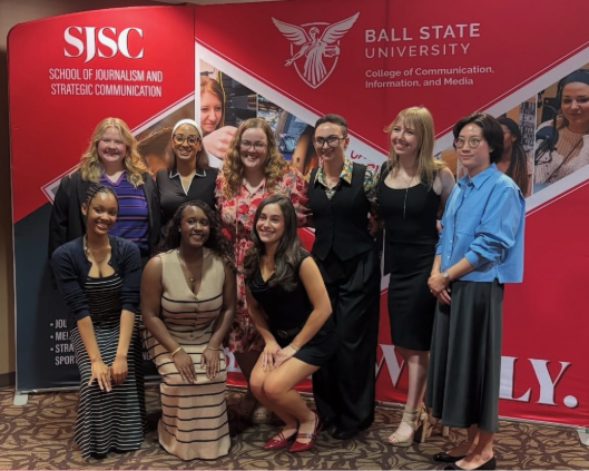 The 2025 Ball State PRSSA Chapter poses in front of a SJSC backdrop that is red and features photos. 