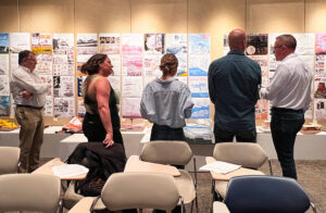 4 jurors along with Ron Menze stand in a line in front of a bulletin board with students project boards and models displayed for judging.