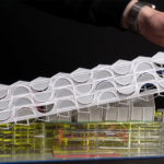 Two hands hold a white, 3D-printed architectural model of a building with curved panels, resting on a yellow translucent base.