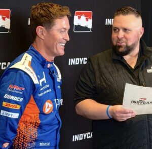 A person in a blue and orange racing suit smiles while speaking with another person holding a document. They stand in front of a black backdrop featuring INDYCAR Radio logos during a media interview or event.